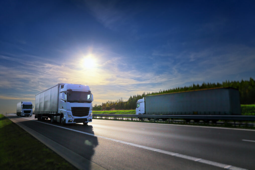 Three large white semi-trucks drive on a highway under a blue sky with the sun shining brightly, bordered by trees and green grass.