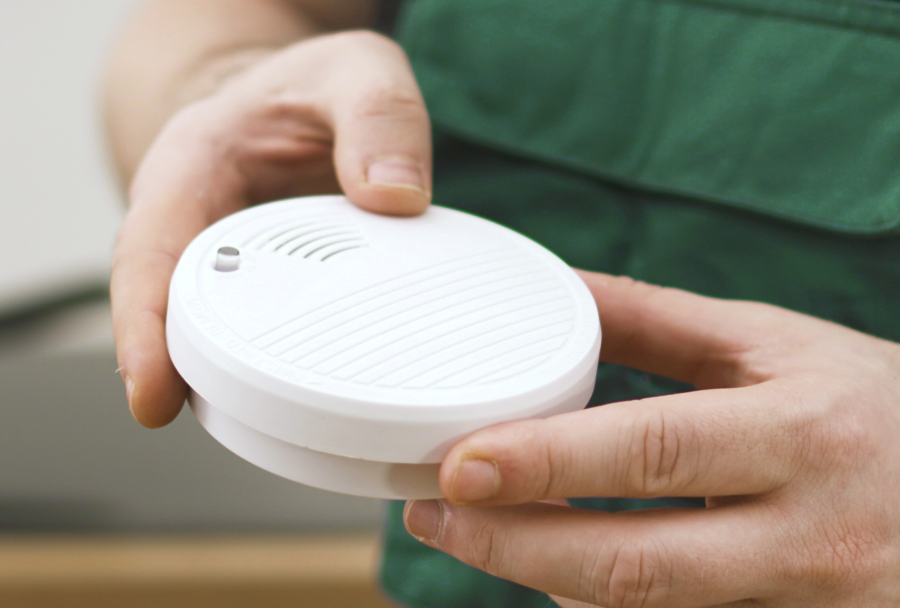 A person in a green shirt holds a white, round smoke detector in both hands.