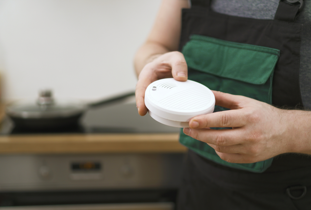 A person in a work apron holds a round smoke detector in a kitchen, preparing to install it.