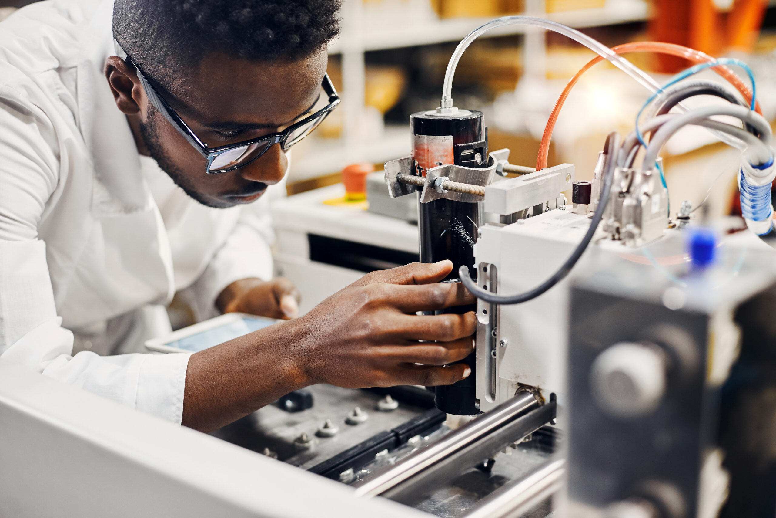 A man wearing glasses and a lab coat adjusts a component on industrial machinery in a laboratory or manufacturing setting.