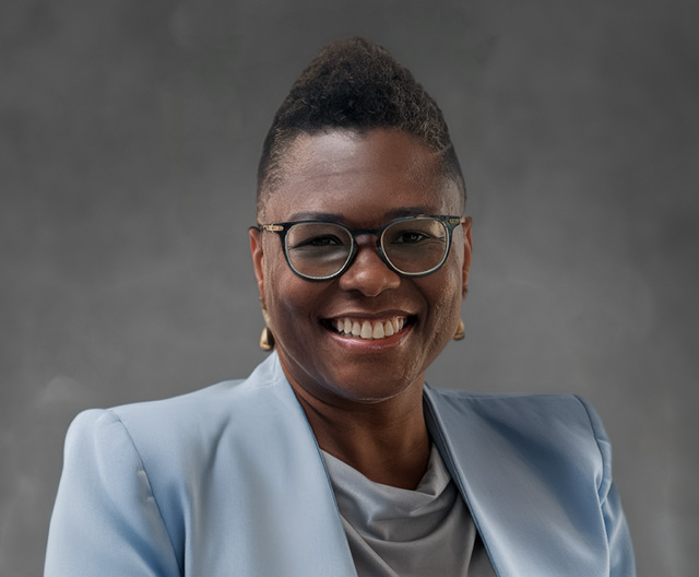 A smiling person wearing glasses, a light blue blazer, and a gray top poses against a plain, dark gray background.