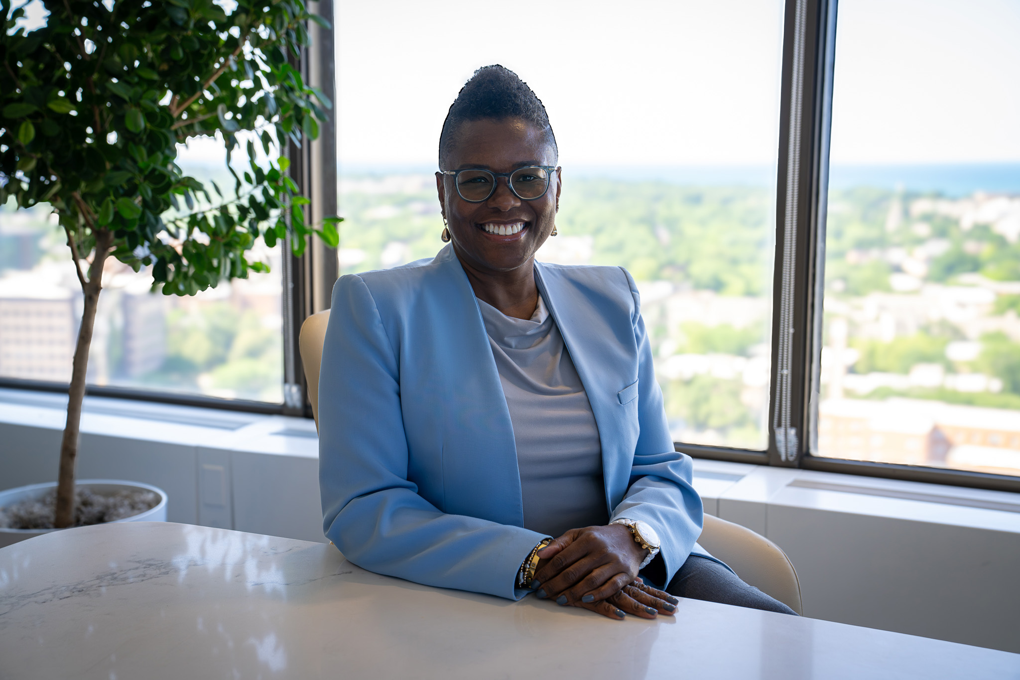 A woman wearing glasses and a light blue blazer sits at a desk, smiling, with large windows and a cityscape in the background. A potted plant is nearby.