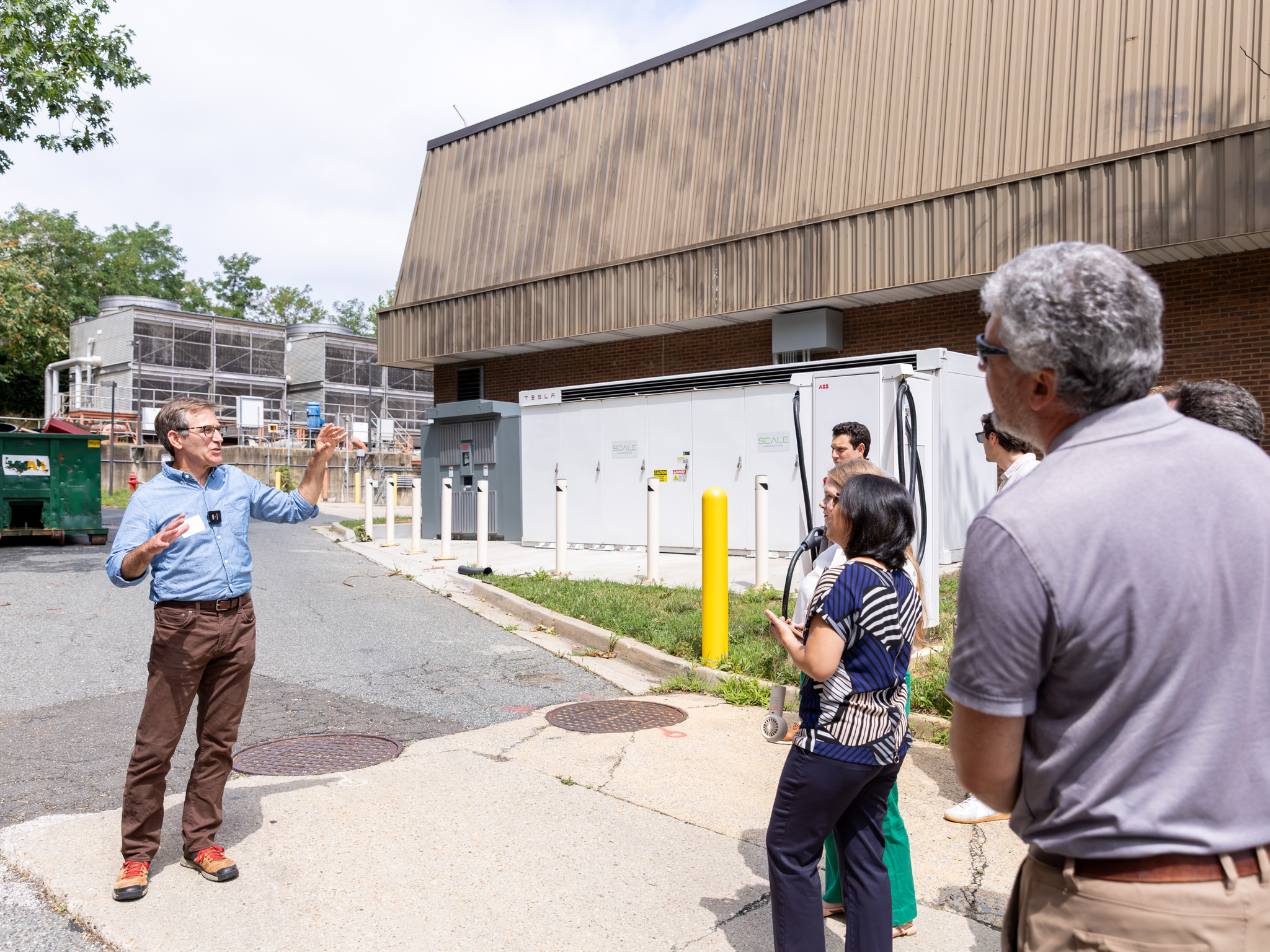 A man stands outside a building, gesturing as he speaks to a small group of people who are listening attentively. The setting includes utility boxes and a parking lot.