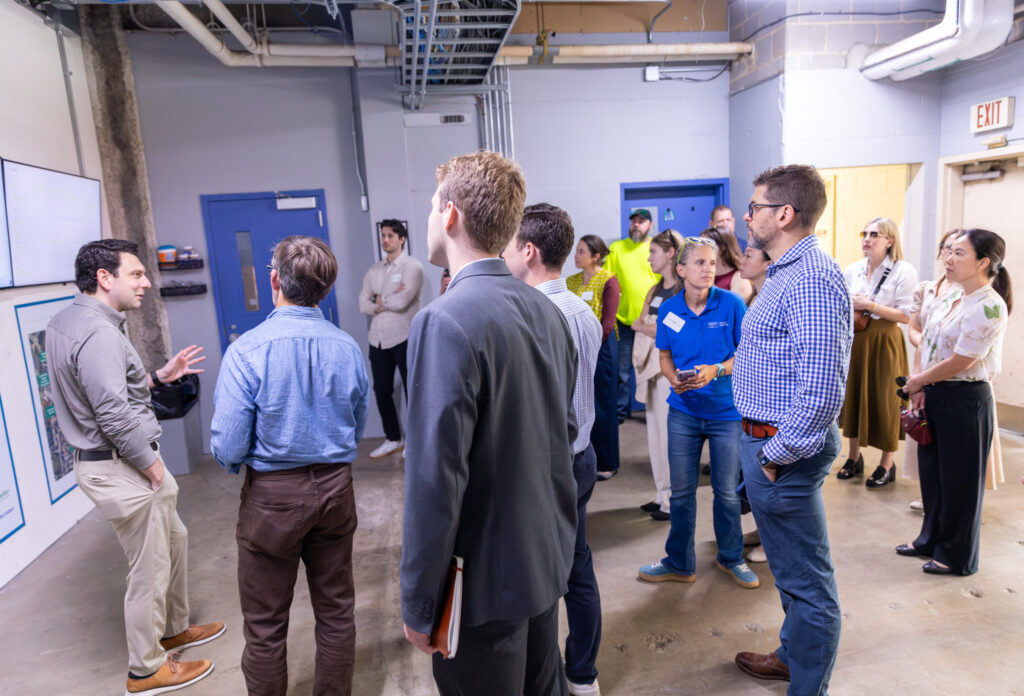 A group of people stands in an industrial room, listening to a man giving a presentation near screens and diagrams on the wall. Some attendees hold notebooks, and a few wear name tags or bright yellow shirts.
