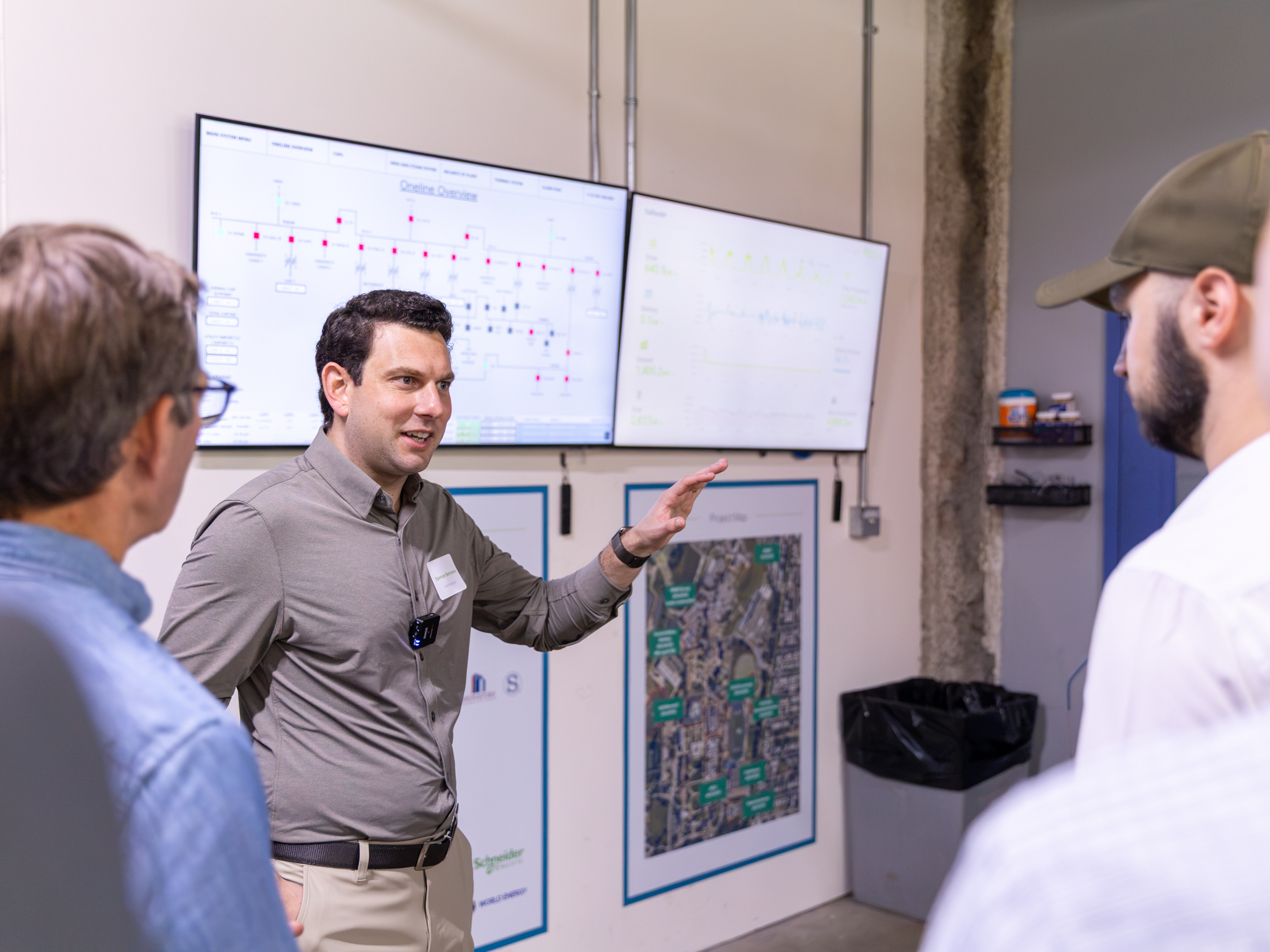 A man gestures while speaking to two others in a room with large screens displaying charts and maps on the wall behind them.