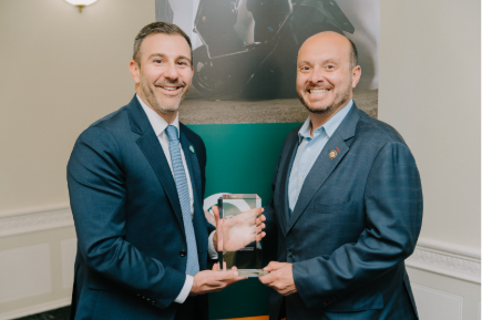 Two men in suits smiling and holding a glass award together, standing indoors in front of a banner.