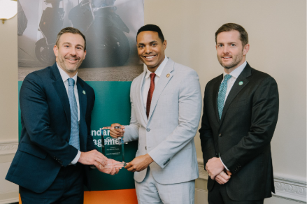 Three men in suits pose indoors in front of a colorful banner; two are shaking hands while one holds an award. All are smiling at the camera.