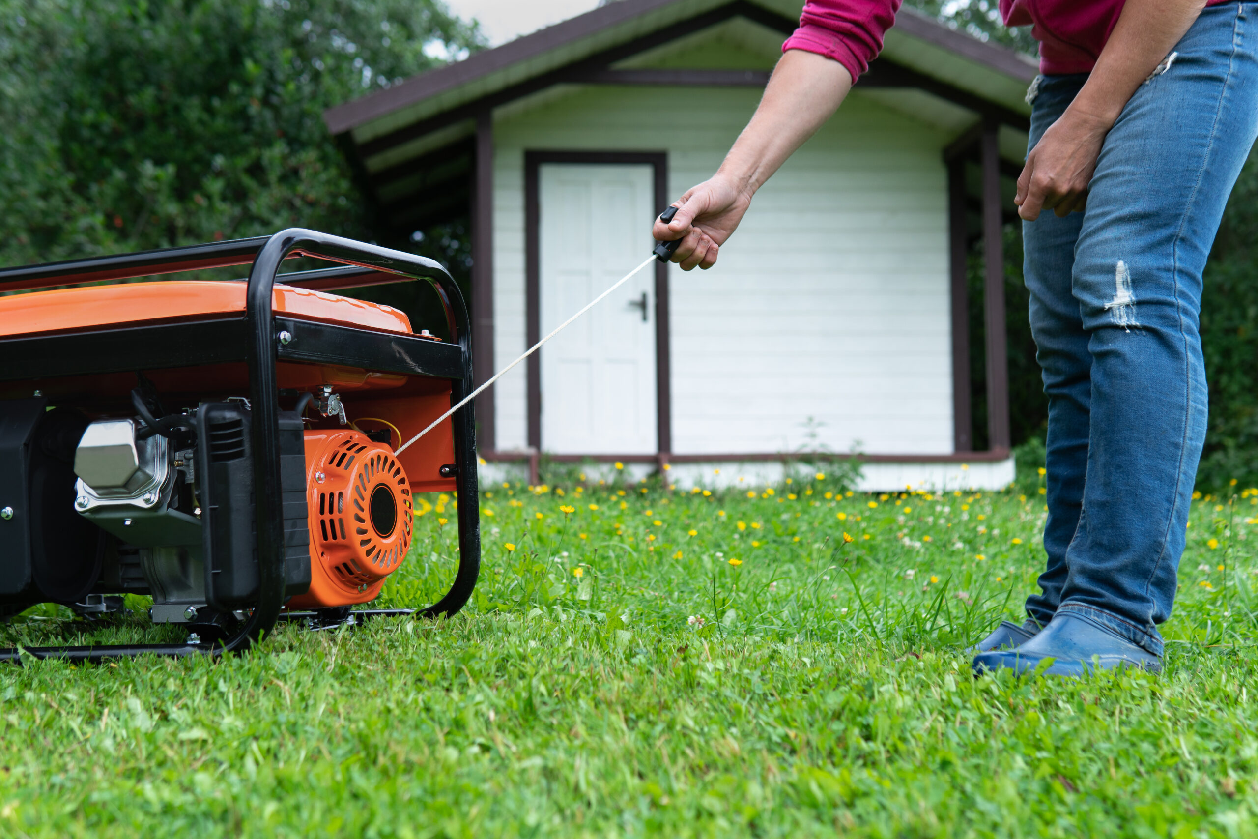 A person pulls the starter cord of an orange portable generator on grass, with a small white shed in the background.