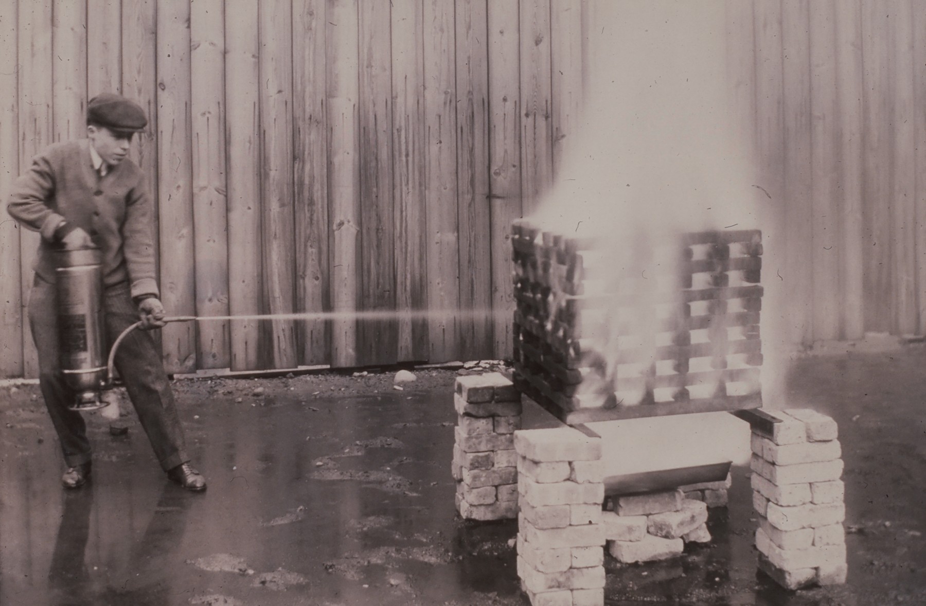 A man in a cap and coat uses a fire extinguisher on a burning stack of wooden crates elevated on brick supports, in front of a wooden fence.