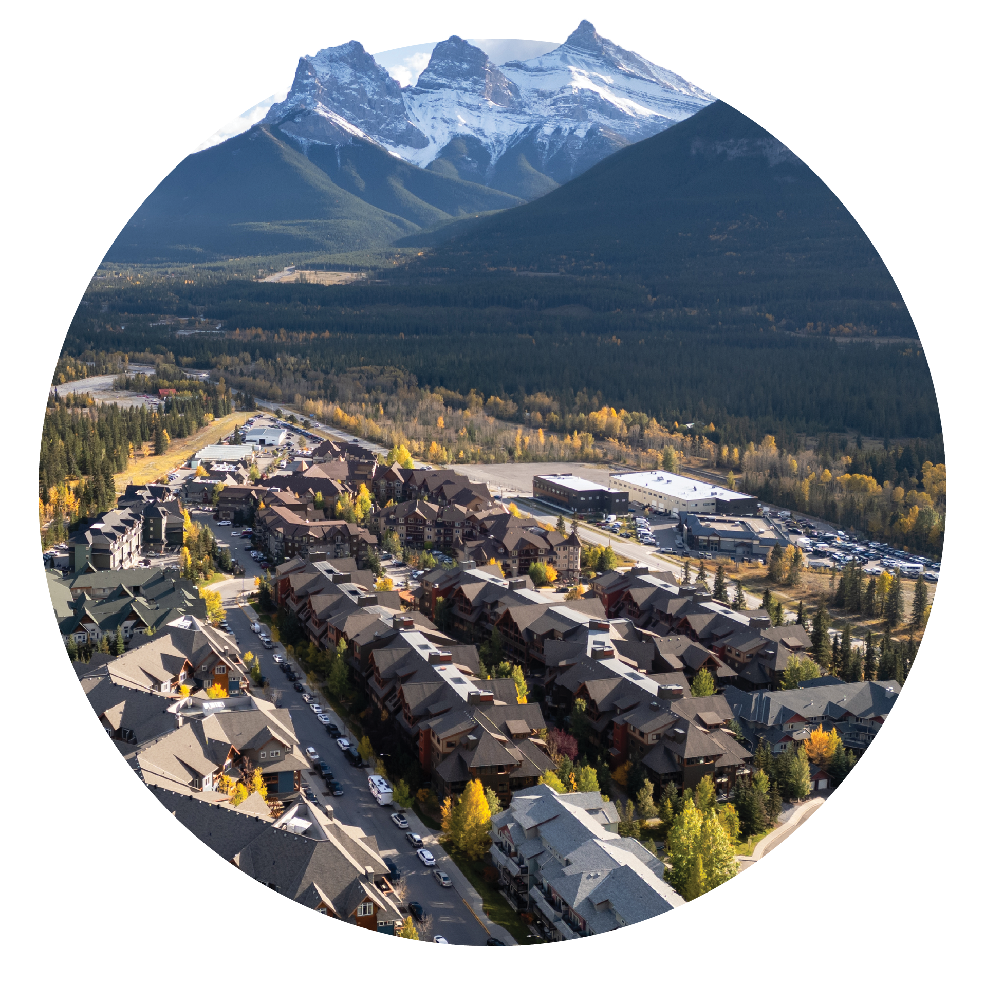 Aerial view of a small town with houses and trees in autumn, with tall, snow-capped mountains in the background under a clear sky.