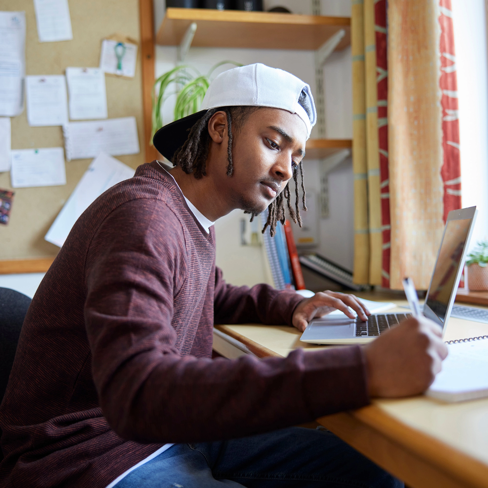 Young man wearing a backward cap sits at a desk, writing in a notebook while looking at a laptop screen in a home office or study room.