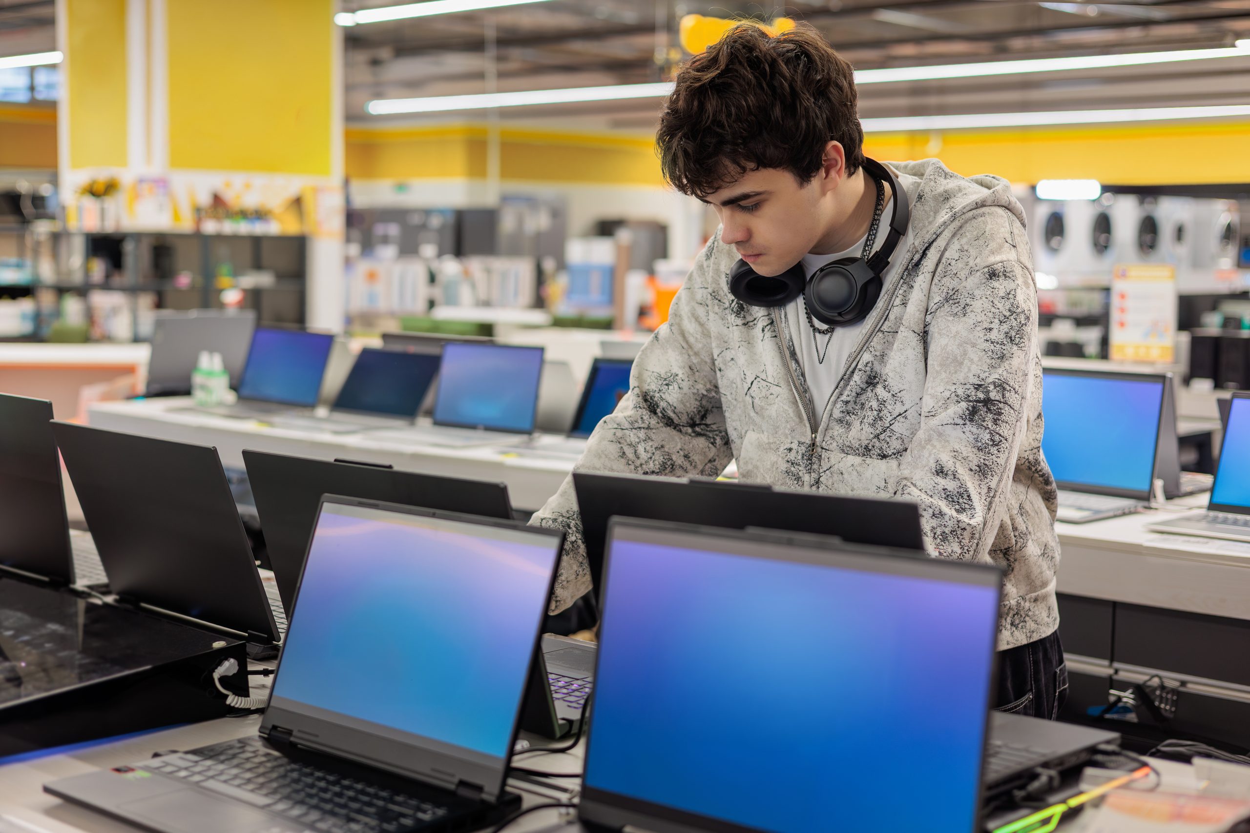 A young man with headphones around his neck examines a laptop in an electronics store, surrounded by several open laptops on display.