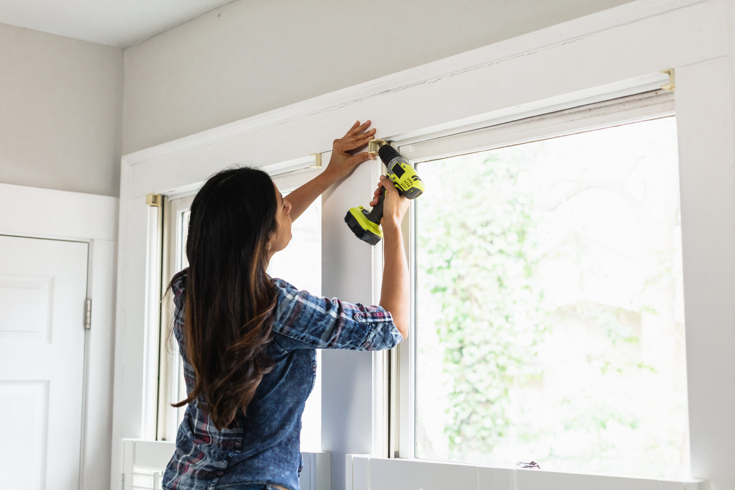 A woman uses a power drill to install or adjust a window treatment above a large window in a bright room.
