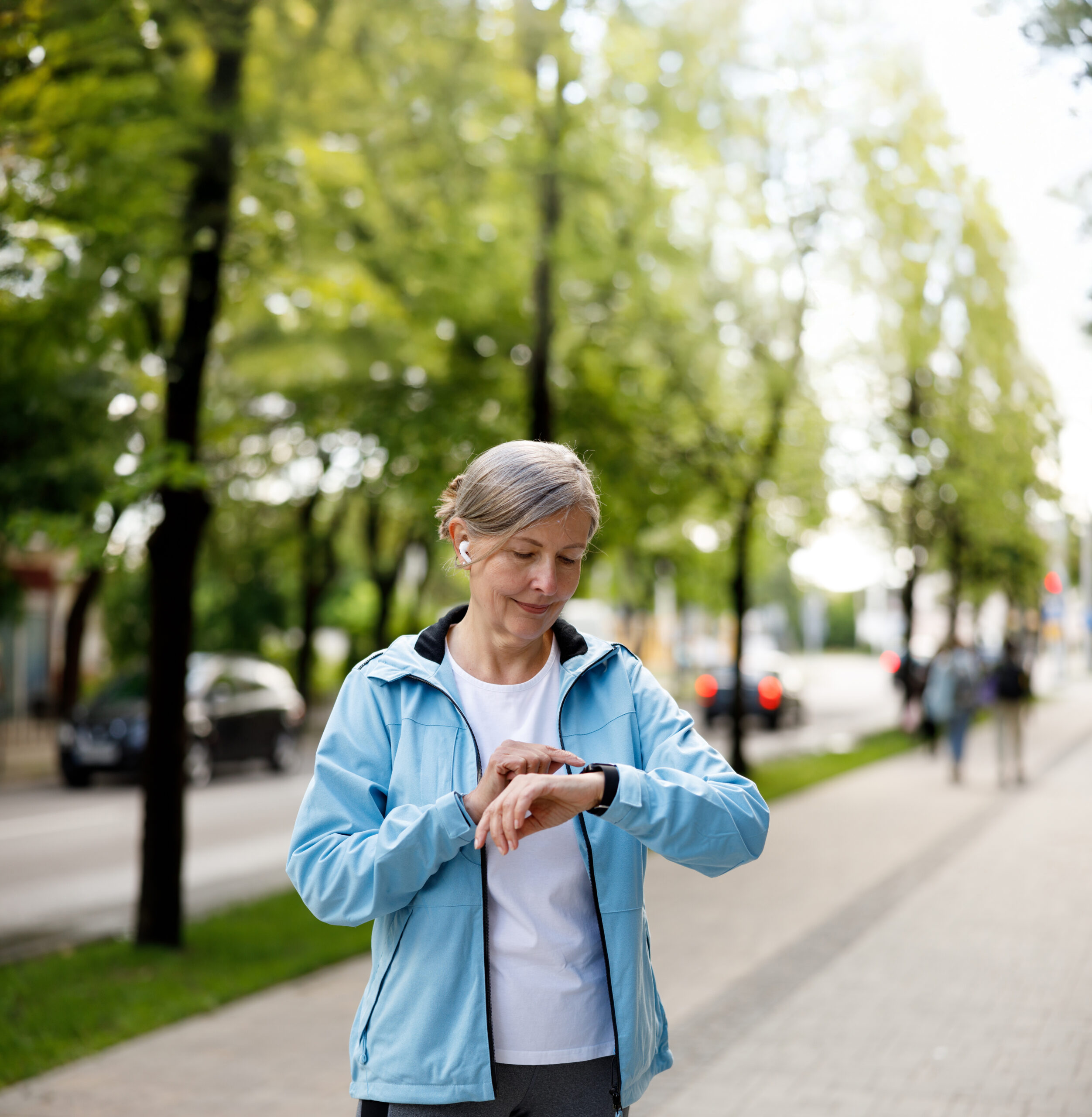 An older woman in a light blue jacket stands on a tree-lined sidewalk, looking at her smartwatch, with cars and people in the blurry background.