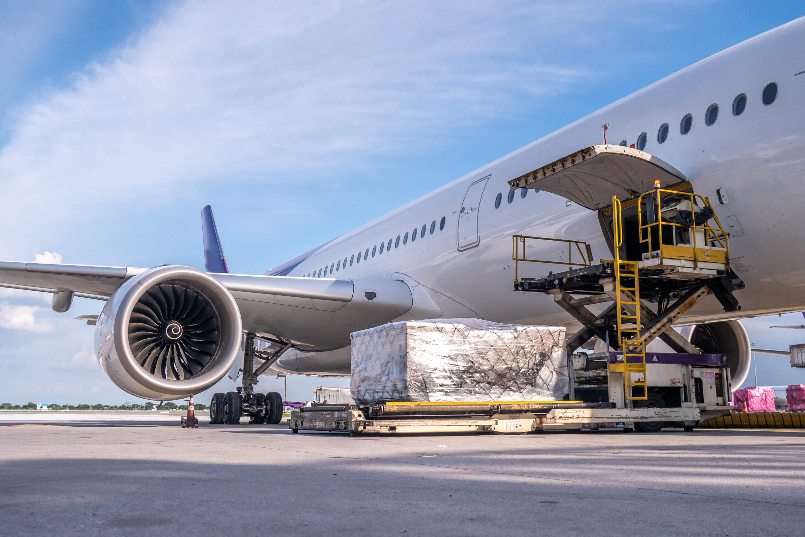 A cargo loader is lifting freight into the open cargo hold of a large commercial airplane on an airport tarmac under a blue sky.