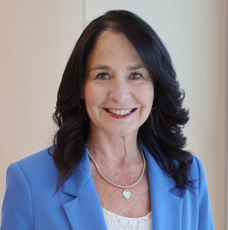 A woman with long, dark hair wearing a blue blazer, white top, silver necklace, and hoop earrings smiles at the camera in a well-lit indoor setting.