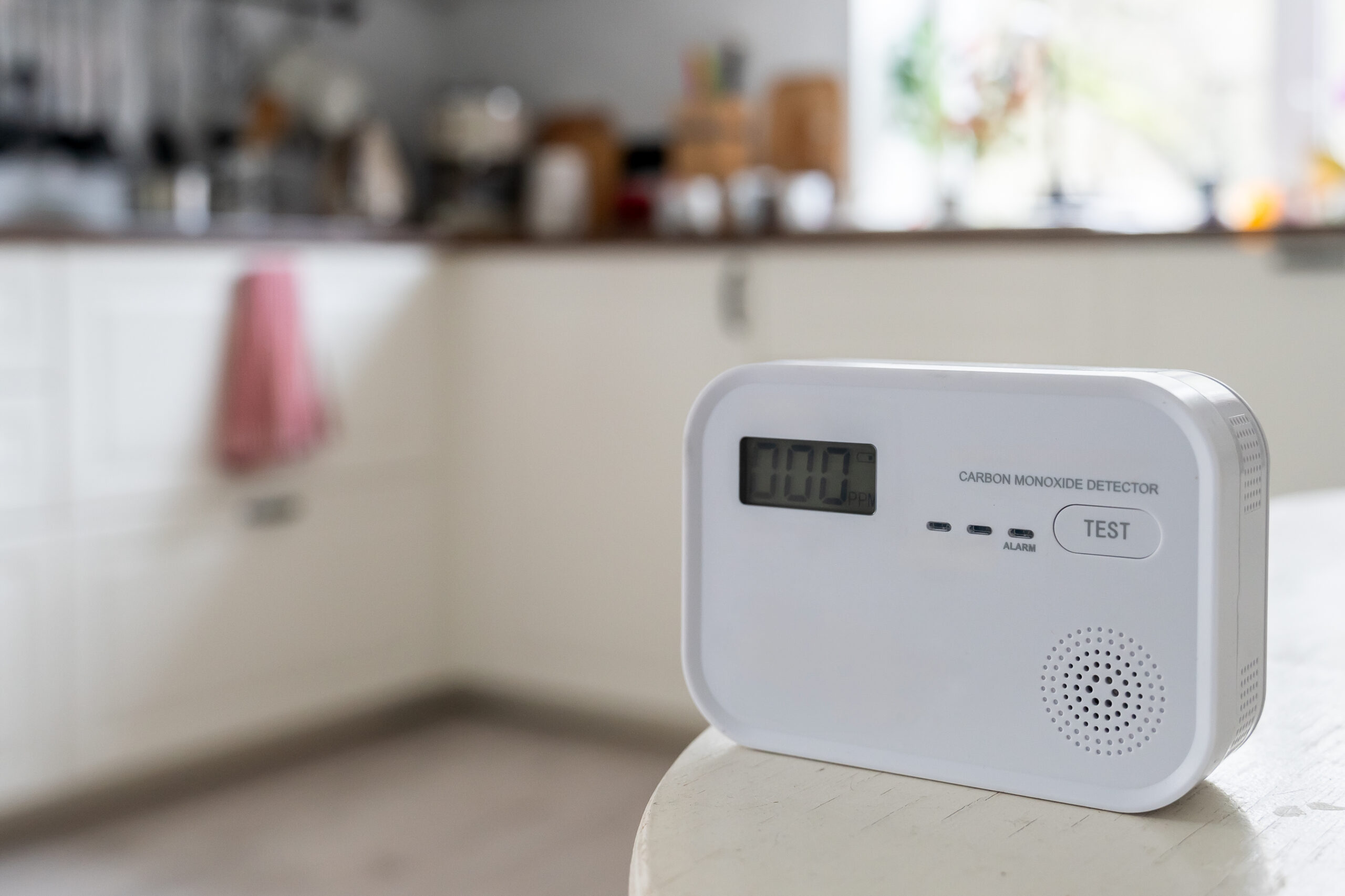 A digital carbon monoxide detector sits on a white table in a bright kitchen, displaying a reading of 0.00 on its screen. The background shows blurred kitchen counters and shelves.