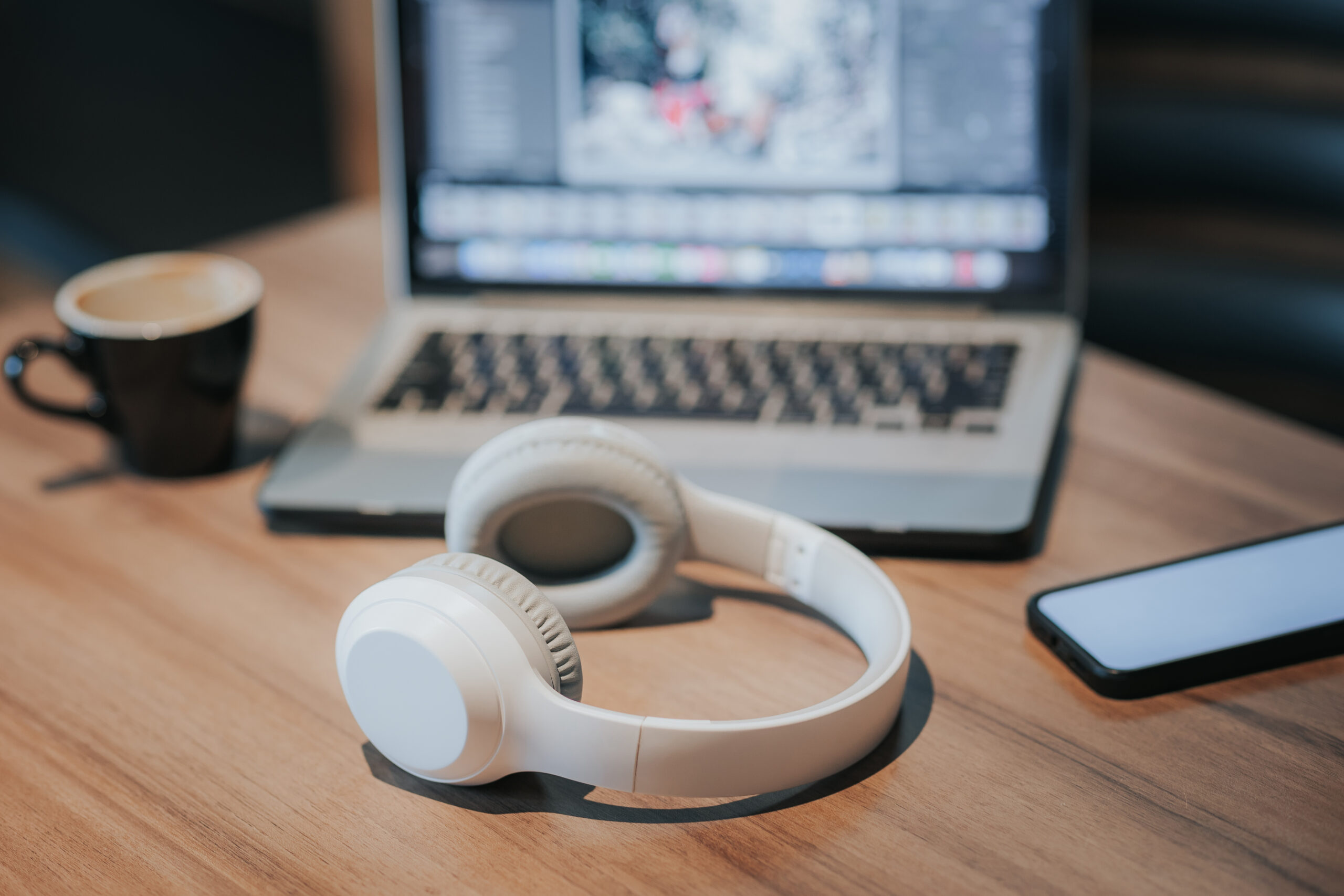 White headphones rest on a wooden table beside a smartphone, a cup of coffee, and an open laptop displaying a blurred screen in the background.