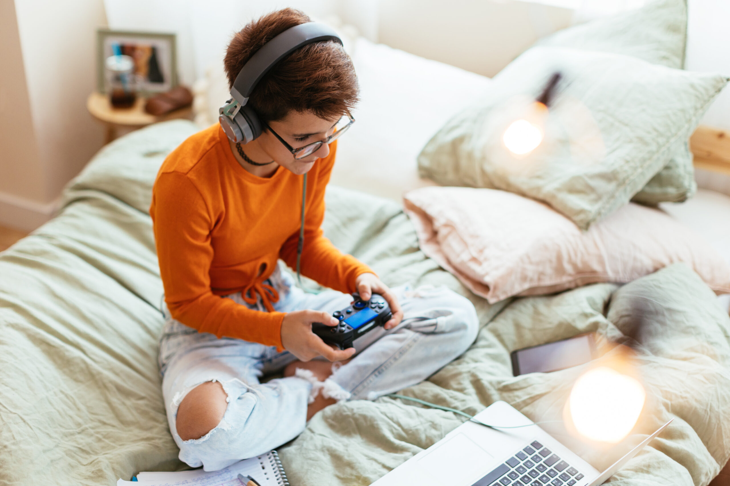 Person wearing headphones and an orange shirt sits cross-legged on a bed, playing a video game with a controller. A laptop, smartphone, and notebook are nearby.
