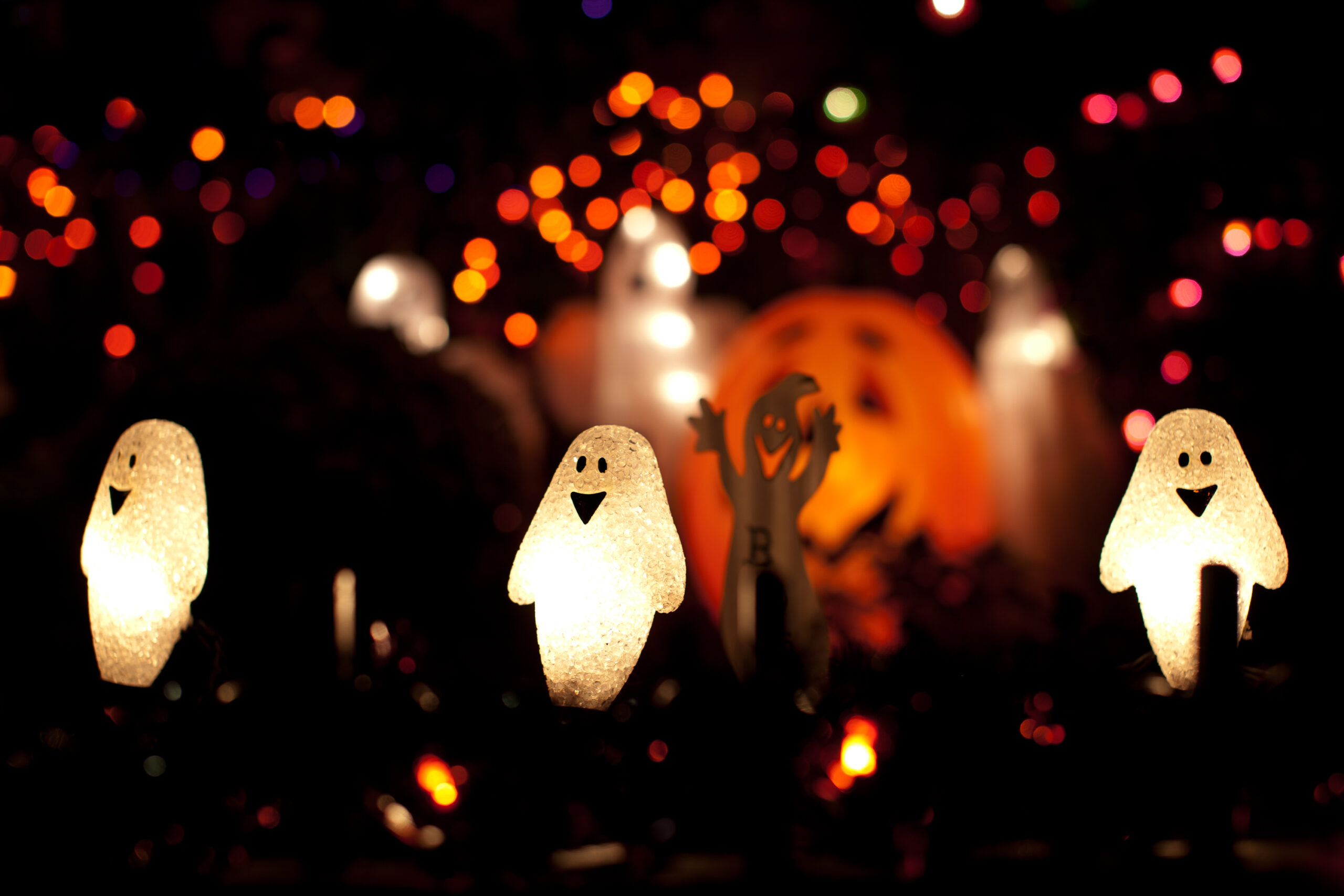 Lighted ghost-shaped Halloween string lights against a dark background with blurred orange bokeh lights.