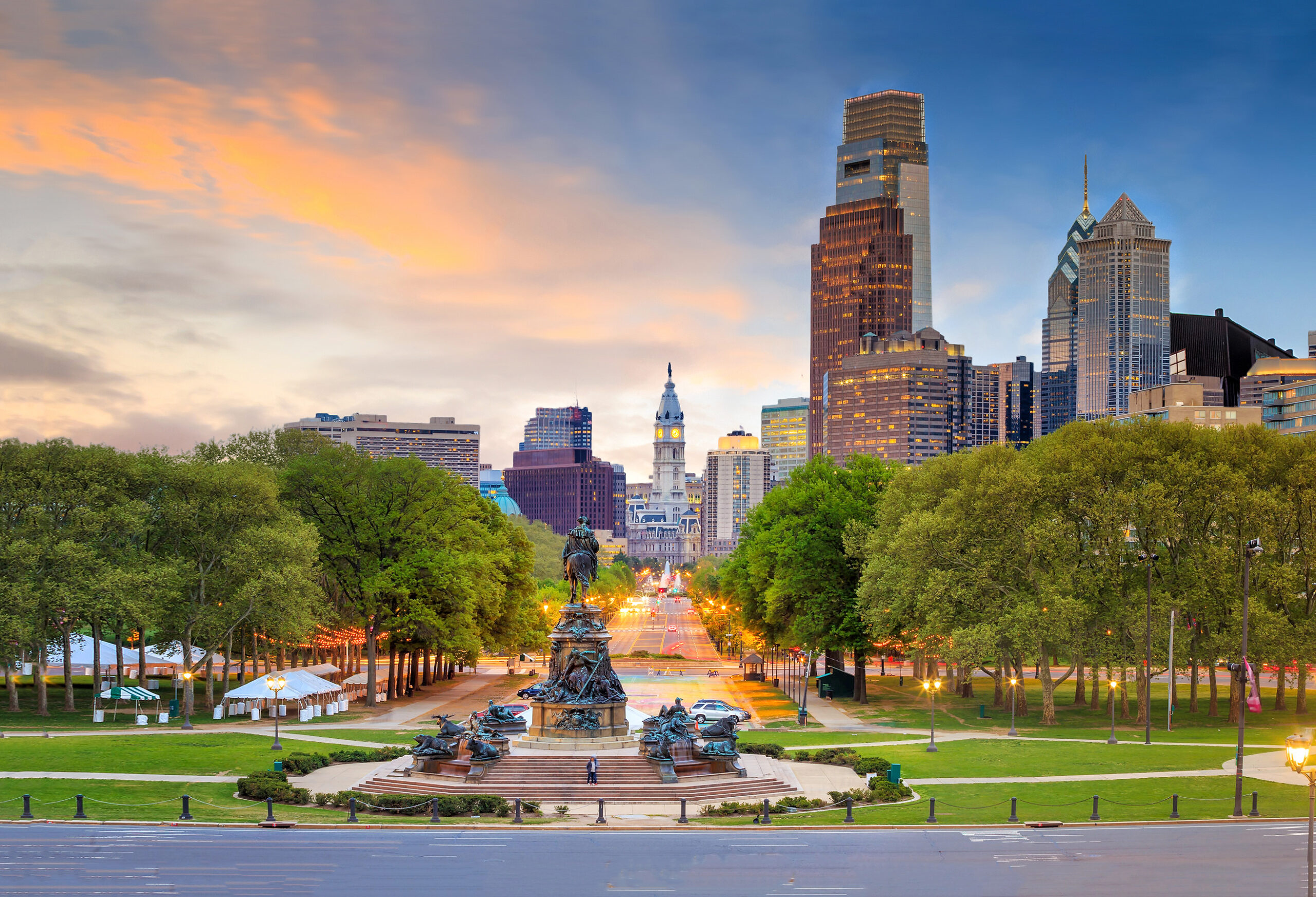 The Philadelphia skyline at dusk, centered on the Washington Monument fountain in front of tree-lined park paths and City Hall.
