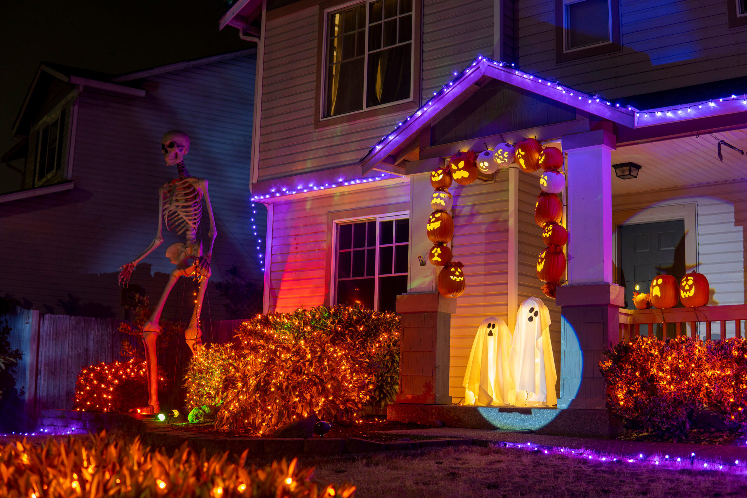A house decorated for Halloween with a giant skeleton figure, glowing pumpkin lanterns lining the porch, and two lit ghost decorations on the front step.